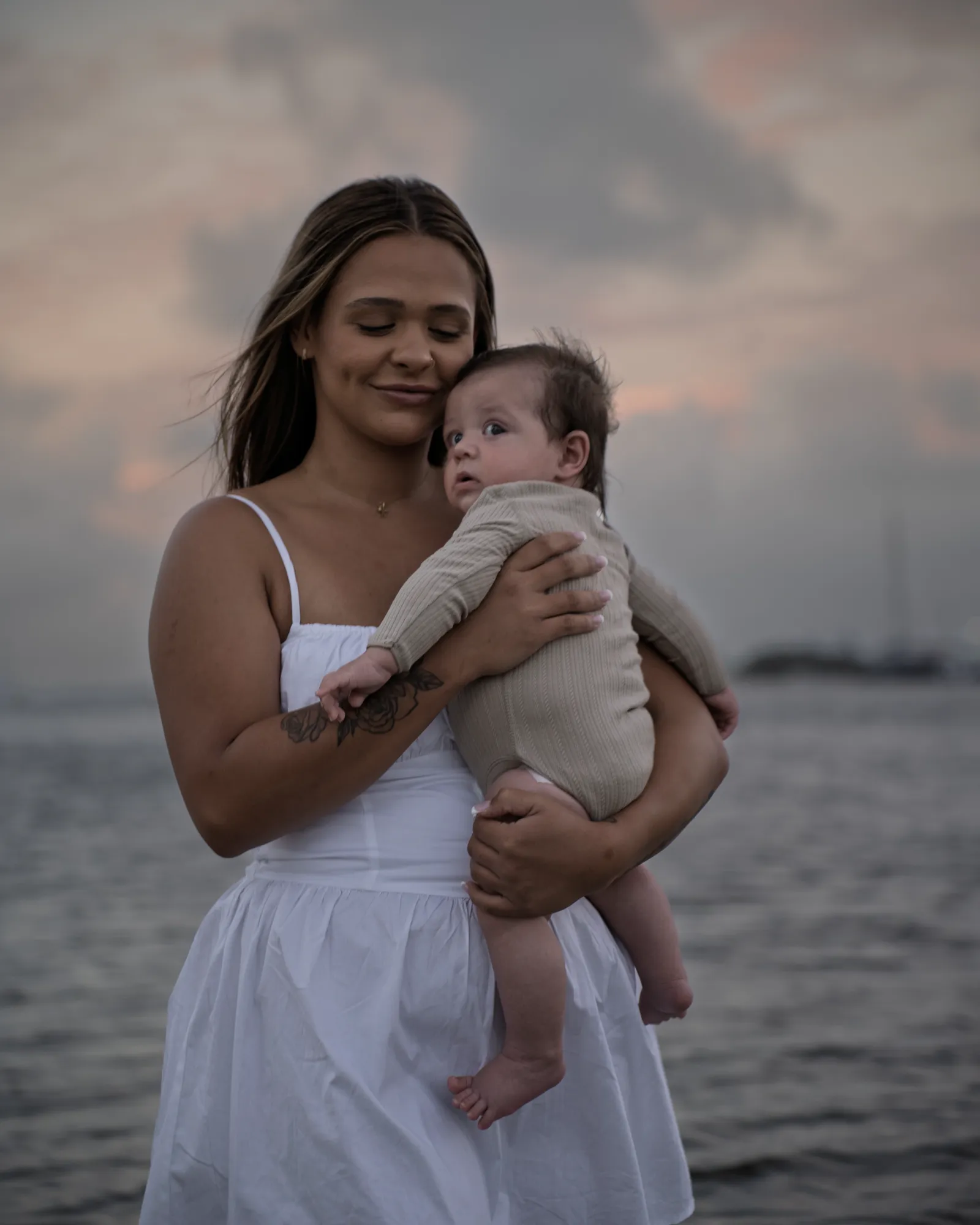 A mother in a white dress holding her baby cheek to cheek, water and sunset cloud behind