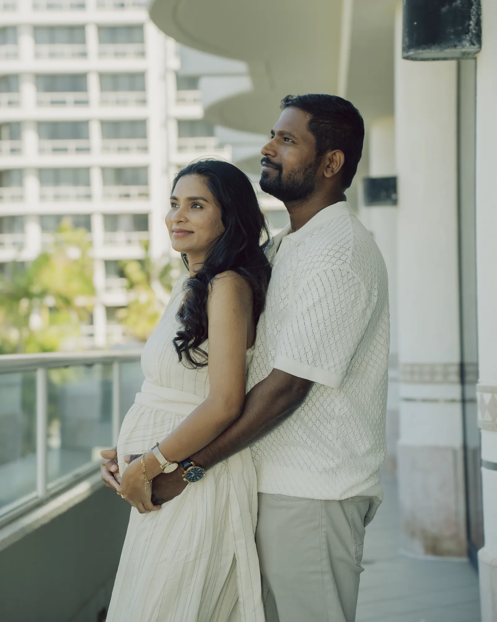 An expecting couple standing together on a balcony, hands on the bump