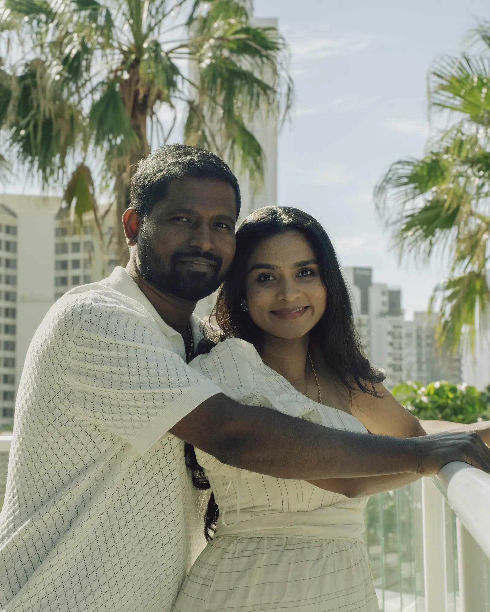 An expecting couple facing camera with palms and high-rises behind
