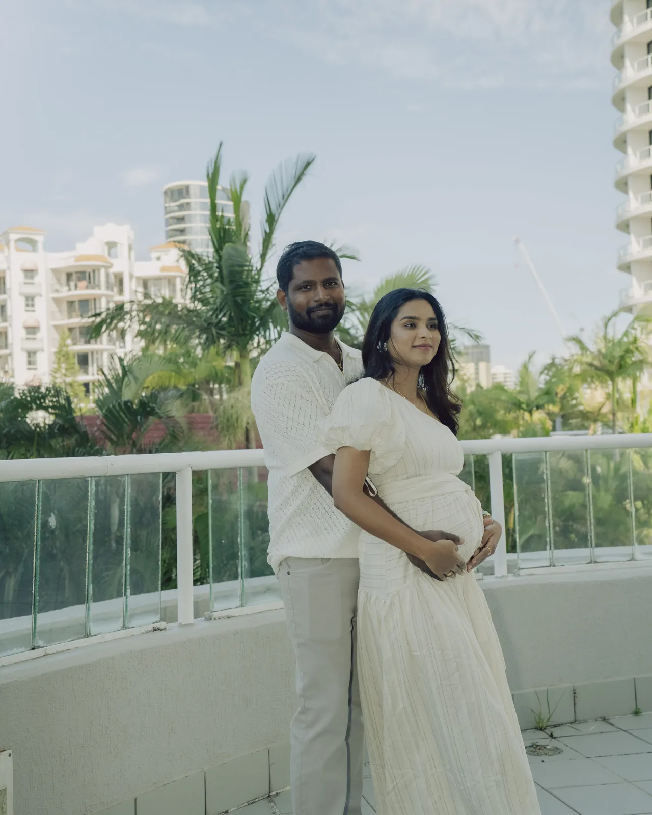 An expecting couple at the balcony rail with palms and towers behind